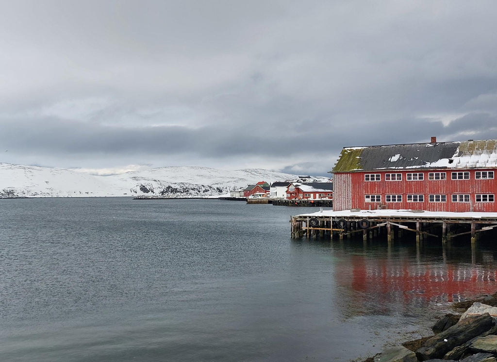 Mehamn havn i Finnmark med røde sjøhus på kaia, snødekte fjell og stille vinterlig sjø