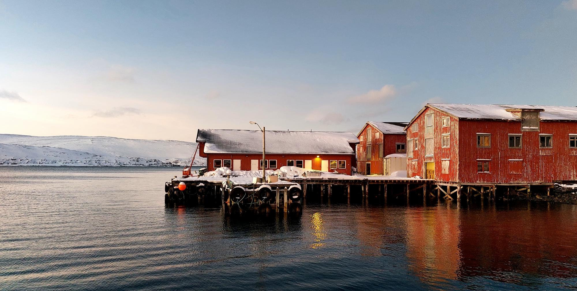 Kaia i Mehamn i Finnmark med røde sjøhus og stille vann i mykt kveldlys
